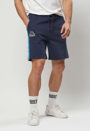 Homme portant un short Kappa bleu marine avec une bande latérale bleue, des chaussettes blanches Kappa, des baskets blanches et un t-shirt blanc, debout les mains le long du corps.