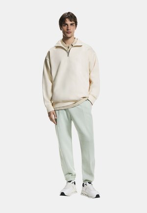 Młody mężczyzna stojący w kremowej bluzie z półzamkiem, beżowej koszulce polo, jasnozielonych spodniach dresowych i białych butach sportowych na białym tle.