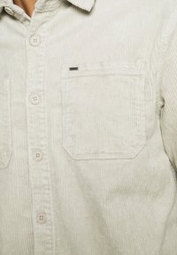 Camisa beige de pana con botones frontales y un bolsillo cuadrado en el pecho que presenta una pequeña etiqueta metálica de la marca en el borde superior del bolsillo.