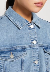 Close-up of a person wearing a light blue denim jacket with silver snap buttons and chest pockets, lips and jawline visible.