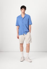 Camisa de manga corta azul clara con un solo bolsillo en el pecho, combinada con pantalones cortos beige. El modelo lleva zapatillas blancas y calcetines hasta el tobillo.