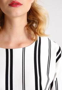 Vit topp med svarta vertikala ränder, med en bred halsringning och textilstruktur. Håret är vågigt, med läpparna målade i en glansig nyans.