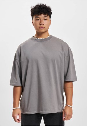 Młody mężczyzna o kręconych włosach, ubrany w luźny szary t-shirt, czarne spodnie, naszyjnik z muszelek oraz dwie bransoletki z muszelek, stoi na tle jednolitego białego tła.