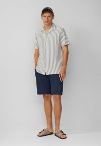Korte mouwen gestreepte shirt in lichtroom en blauw, gecombineerd met marineblauwe shorts. Het model draagt bruine slippers met een banddetail.