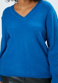 Blauer V-Ausschnitt-Pullover aus weichem, strukturiertem Stoff, mit kleinen glitzernden Akzenten und gerippten Bündchen und Saum.