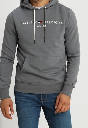 Mężczyzna w szarej bluzie z kapturem Tommy Hilfiger z białymi troczkami i logo, w połączeniu z niebieskimi jeansami, stojący na jednolitym tle.