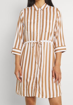 Kvinde iført knælange kjole med knapper, brun og hvide lodrette striber, lange ærmer, to brystlommer og bindebånd i taljen.