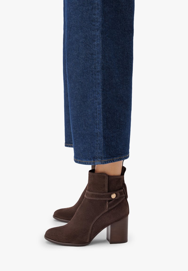 Botine din suede maro, cu vârf rotunjit, un design curbat și toc bloc maro, având o curea decorativă și un accent cu buton.