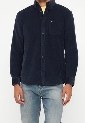 Hombre con una camisa de pana azul marino oscuro con bolsillo en el pecho y vaqueros azul claro, de pie frente a un fondo liso.