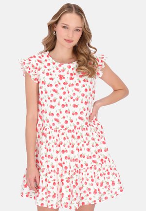 Nuori nainen, jolla on laineikkaat hiukset, pukeutunut valkoiseen mekkoon, jossa on punainen kirsikkakuosi ja röyhelöidyt lyhyet hihat, poseeraa käsi vyötäröllä.