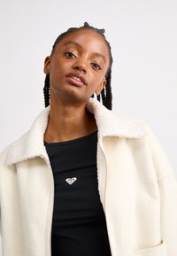 Veste en fausse fourrure couleur crème avec un col doux, fermeture éclair sur le devant et poches latérales, portée sur un top noir à côtes avec un petit logo en forme de cœur.