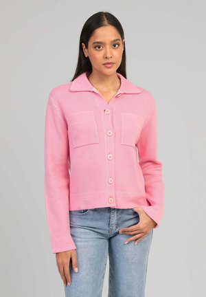 Femme aux longs cheveux foncés portant une veste rose boutonnée avec des poches avant et un jean bleu clair, posant devant un fond uni.