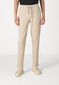 Beige joggebukser lavet af blødt stof, med elastisk taljebånd og snøre, slim fit og opkrøllede manchetter. Sammen med lyse sko.