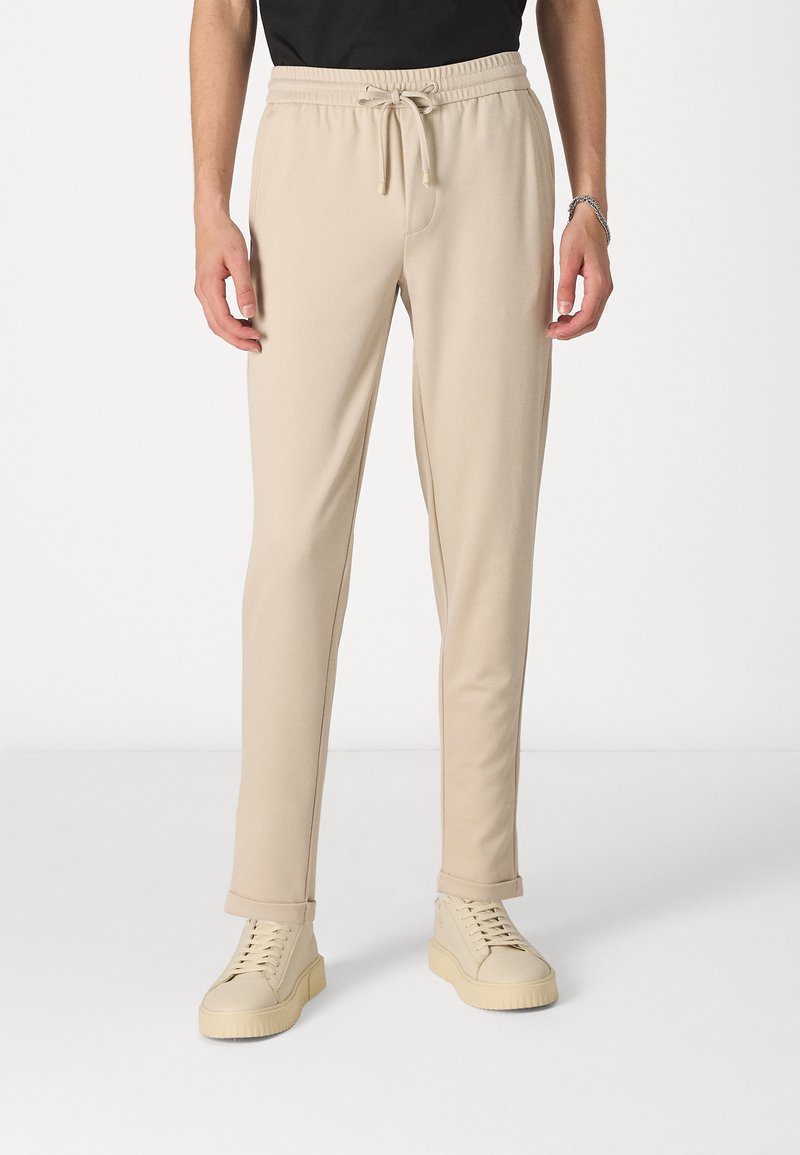 Beige joggebukser lavet af blødt stof, med elastisk taljebånd og snøre, slim fit og opkrøllede manchetter. Sammen med lyse sko.