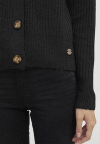 Čierny pletený kardigán s rebrovouk textúrou, hnedými dekoratívnymi gombíkmi a surovými okrajmi na lemoch a rukávoch. Nosený s čiernymi nohavicami.