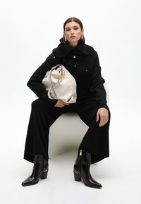 Abrigo de punto negro con un cuello alto, acentos de hardware dorado, pantalones anchos negros y botas de tobillo negras brillantes, sosteniendo un bolso beige con un cierre dorado.