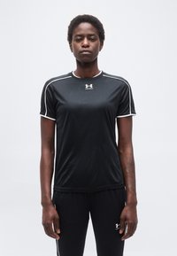 Αθλητικό άτομο που στέκεται όρθιο φορώντας μαύρα αθλητικά ρούχα Under Armour με λευκή διακόσμηση στα κοντά μανίκια και το παντελόνι, πάνω σε απλό φόντο.