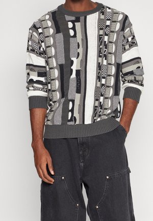 Man in een meerkleurige grijs, zwart en witte gebreide trui met ribgebreide mouwen en donkergrijze broeken met grote voorzakken.