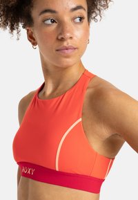 Sportbehå i stark orange med ett rött band. Har en hög halsringning, en smidig design och kontrasterande sömmar. Mjuk, elastisk tyg.