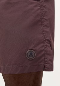 Șorturi de înot burgundy, fabricate dintr-un material ușor, cu o textură netedă. Prezintă un patch rotund cu un logo ce include un craniu și două oase încrucișate.