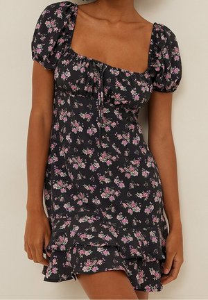 Sort blomsterkjole med små blomster i pink og hvid, pufærmer, firkantet udskæring og lagdelt flæsekant, båret af en person med mellemtonet hud.