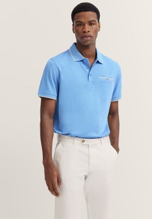 Lichtblauw poloshirt met een klein voorzakje en witte afwerking. Draagt met witte broek, voorzien van knoopsluiting en zijzakken.