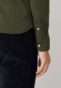 Camisa de algodón color verde oliva con un dobladillo recto, puños abotonados y detalles de costura visibles. Llevada con pantalones de pana oscuros.