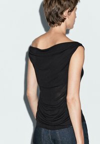 Svart off-shoulder topp med draperad halsringning och samlade sidor, tillverkad av ett slätt och lätt tyg. Bärs med blå jeans.