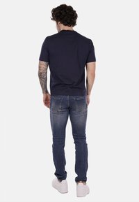 T-shirt a maniche corte blu navy, in cotone, con un design aderente. Indossato con jeans azzurri e sneakers bianche, con vista da dietro e tatuaggi.