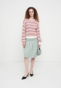 Γυναίκα με σγουρά μαλλιά που φοράει πολύχρωμο ριγέ πουλόβερ, φούστα σε ανοιχτό μπλε με πιέτες, μαύρες γόβες και κρατάει μια μικρή γκρι τσάντα χειρός.