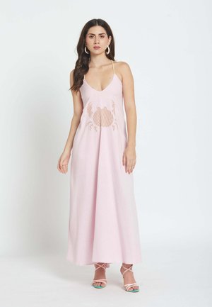 Ženska v brezrokavni svetlo roza maxi obleki z motivom raka na sprednji strani, obuta v roza sandale z naramnicami in z obročastimi uhani, stoji pred belim ozadjem.