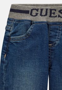 Jean bleu avec une ceinture grise portant l'inscription "GUES" en bleu marine. Le tissu a une texture lisse avec une légère décoloration et des poches avant.