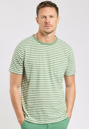 Homme portant un T-shirt rayé vert et blanc et un pantalon vert, debout les mains dans les poches, expression neutre, fond clair uni.