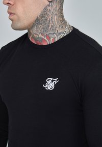 Black long-sleeve shirt with crew neck, featuring a white embroidered logo on the chest. Smooth, stretch fabric without patterns.