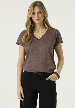 Femme aux longs cheveux blonds porte un haut marron à manches courtes avec un col en V, un pantalon noir, des colliers superposés et des bracelets, debout avec une main dans la poche.