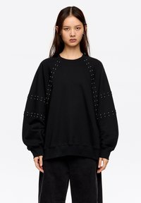 Sudadera negra con detalles de tachuelas en las mangas, corte holgado, escote redondo y puños acanalados. Hecha de tela suave y resistente.