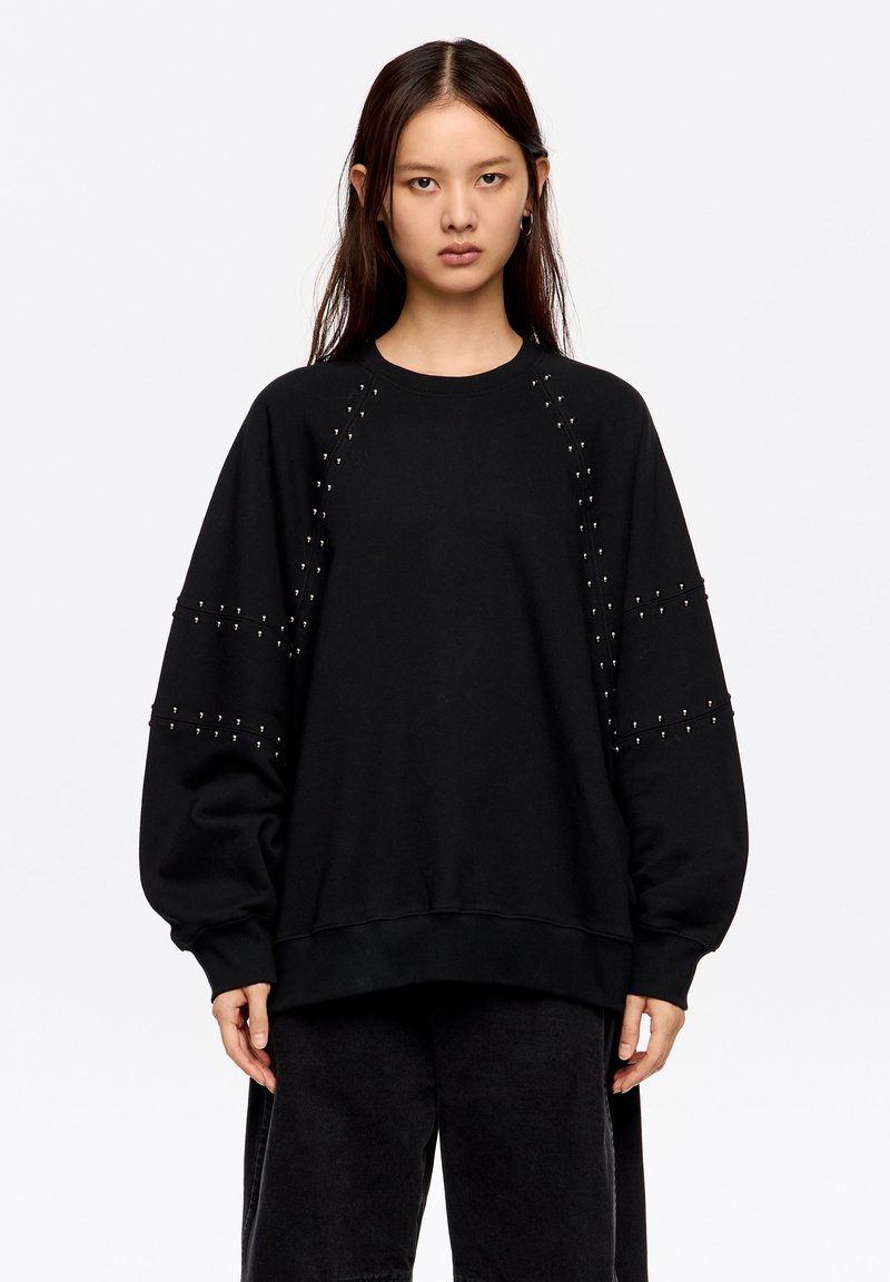Sudadera negra con detalles de tachuelas en las mangas, corte holgado, escote redondo y puños acanalados. Hecha de tela suave y resistente.