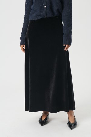 Juodas aksominis ilgis sijonas su lygiu paviršiumi, turintis prigludusią liemenėlę ir laisvą siluetą, derinamas su juodais smailiais bateliais ir tamsiai mėlynu megztiniu.