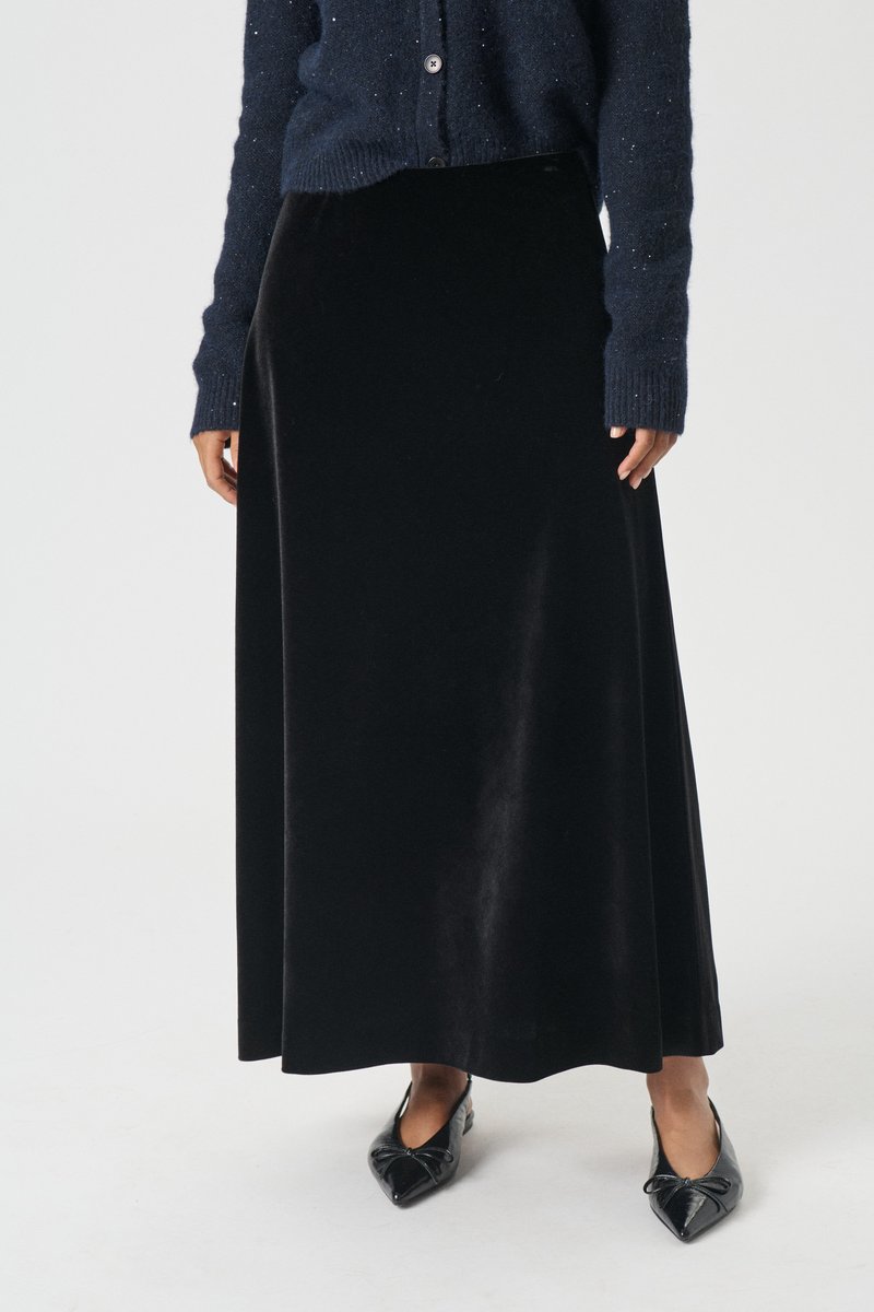 Črna žametna maxi krilo z gladko teksturo, ki ima oprijet pas in tekoč silhuet, v paru s črnimi koničastimi balerinkami in mornarsko modrim kardiganom.
