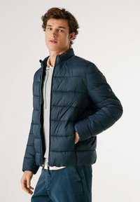 Mornarsko modra puffer jakna z zadrgo, vodoravno prešita tekstura, visok ovratnik in stranske žepe, nosi se čez svetlo sivo majico.