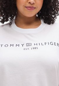 Λευκό βαμβακερό t-shirt με κοντά μανίκια, στο μπροστινό μέρος φέρει μεγάλο μαύρο λογότυπο "TOMMY HILFIGER" και το κείμενο "EST. 1985".