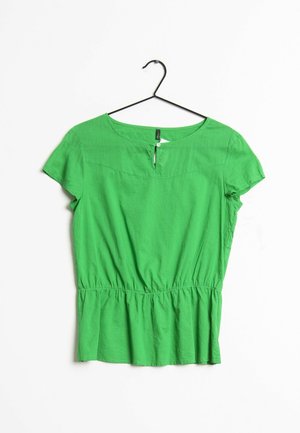 Grøn kortærmet bluse med rund halsudskæring, lille knappedetalje foran, indsamlet talje, hængende på sort bøjle mod hvid baggrund.
