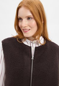 Braune Fleece-Weste mit hohem Kragen und Reißverschluss, ausgestattet mit einer weichen Textur und ärmellosem Design, über einem gestreiften Shirt getragen.
