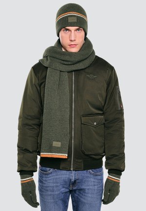 Vyras, vilkintis žalią megztą kepurę, šaliką ir pirštines su oranžinėmis ir baltomis juostelėmis, tamsiai žalią striukę ir mėlynas džinsus, stovintis prieš paprastą foną.