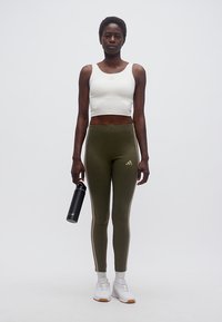 Γυναίκα στέκεται κρατώντας μαύρο παγούρι νερού, φορώντας λευκό κοντό τοπ, ελιάς πράσινο κολάν με ρίγες, λευκές κάλτσες και λευκά αθλητικά παπούτσια σε απλό φόντο.