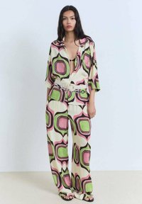 Zweiteiliges Outfit mit einem locker sitzenden Shirt und weit geschnittenen Hosen. Hergestellt aus leichtem Stoff mit einem cremefarbenen Hintergrund und kräftigen geometrischen Mustern in Pink, Grün und Schwarz.
