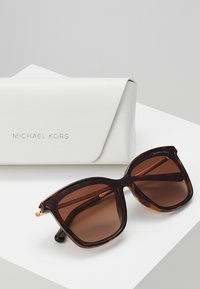 Brown tortoiseshell sunglasses with oversized square lenses and a glossy finish, alongside a white textured hard case marked with "MICHAEL KORS."