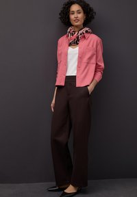 Chemise boutonnée rose clair portée sur un haut blanc, associée à un pantalon large marron foncé ; une écharpe à motifs apporte une touche visuelle. Des chaussures noires complètent la tenue.
