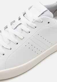 Baskets en cuir blanc avec lacets blancs, petits trous de ventilation perforés et semelle en caoutchouc texturé beige clair.