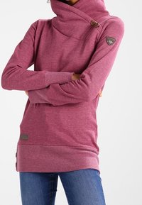 Donna che indossa un maglione rosa a maniche lunghe con collo alto e bottoni vicino al colletto, abbinato a jeans blu, con le braccia incrociate.